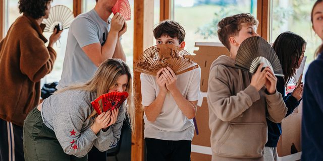 A group of young performers huddle together in a rehearsal room. They hold different coloured fans up to their faces, looking over the top with wide, excitable eyes. They all face different directions. There is a sense of anticipation.