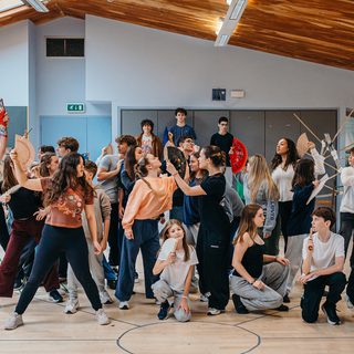 A large group of young performers pose enthusiastically in a rehearsal room. Some of them hold colourful fans up to their faces. They all look in different directions.