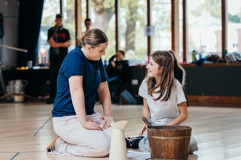 Two young performers sit on the floor of a rehearsal room. A younger girl admiringly looks up at an older girl with a smile. The older girl looks back at her with a gentle, kind smile. There is a wooden bucket and a metal jug on the floor in front of them. Other young performers watch on in the background, out of focus.
