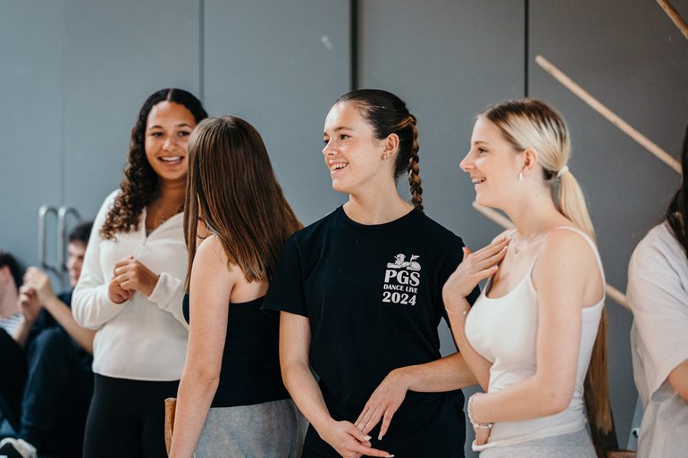 Four young performers standing in a rehearsal room, photographed from the side. They are talking and laughing, looking relaxed, enjoying themselves.