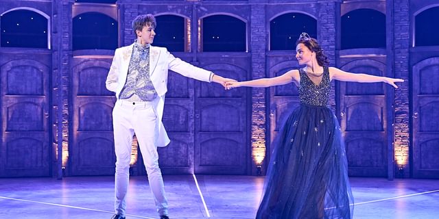 Two young performers dance onstage. They hold their hands in the centre, outstretched and look at one another intently. They look a little shy but happy to be together. The performer on the left wears a white tail suit with a blue floral silk waistcoat and tie. The performer on the right wears a stunning deep blue dress with an embellished bodice and layered tulle skirt scattered with silver flowers with a silver crown in their dark hair, tied back in a bun.