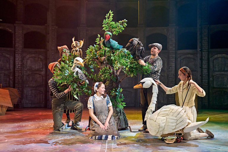 A young performer sits onstage in front of a tree, surrounded by charming, colourful bird puppets: a swan, an owl, a parakeet, a jackdaw and a gull. She looks at the swan with a loving smile. She wears a light brown cotton apron, striped blue and cream skirt, a floral white blouse and a denim waistcoat with hair tied up in a scarf.