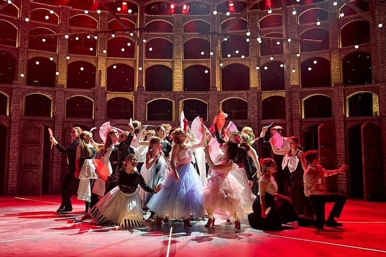 A striking scene onstage with young performers posing in a tableau holding fans, dressed in party costumes. They pose in a variety of different positions and levels, looking up in different directions. The stage is lit in a vibrant red with white light bulbs hanging from the ceiling.