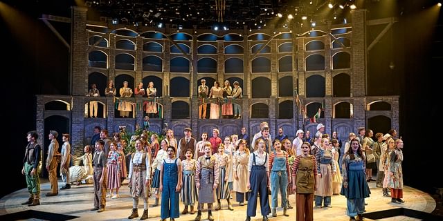 The stage is full of young performers singing enthusiastically. They are all having an amazing time performing. They all wear different costumes like they are from a country village, doing a variety of jobs. They look out to the audience, wide eyed. In the background is a curved wall with lots of windows in, giving the sense of a village behind them.