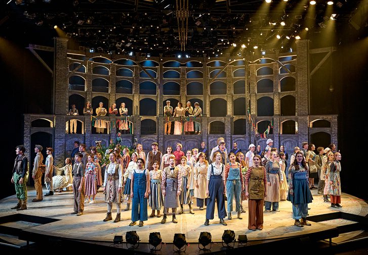 The stage is full of young performers singing enthusiastically. They are all having an amazing time performing. They all wear different costumes like they are from a country village, doing a variety of jobs. They look out to the audience, wide eyed. In the background is a curved wall with lots of windows in, giving the sense of a village behind them.