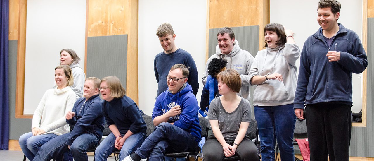 The Friday Company are pictured sitting and standing on the stage in the Pimlott Building. They are smiling and laughing.