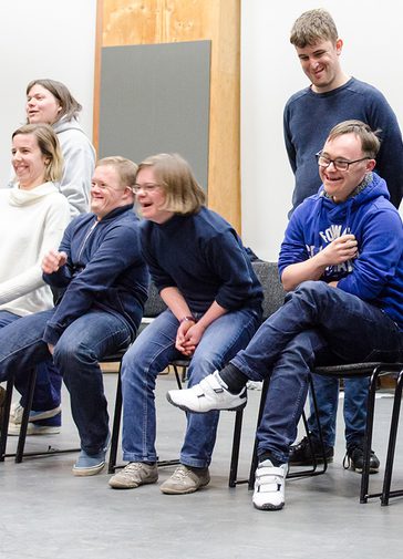 The Friday Company are pictured sitting and standing on the stage in the Pimlott Building. They are smiling and laughing.