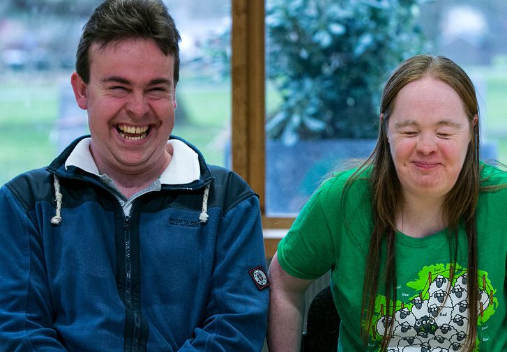 A man and woman are sat down in the Pimlott building, smiling and laughing. He is on the left and wearing a blue coat. She is on the right and is wearing a green t-shirt with sheep on it. Behind them you can see Oaklands Park.