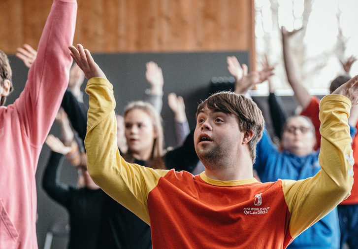 A group of young people wearing colourful clothes are dancing. Their hands are raised in the air above their heads, and they are smiling.