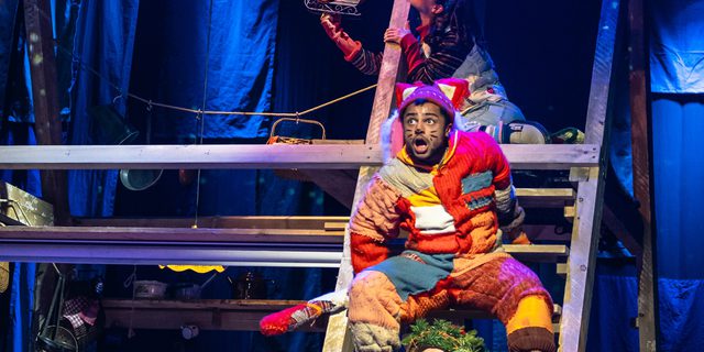 Three performers sit on a ladder that forms part of a wooden a-frame structure. The performer at the bottom is dressed as a Christmas tree, above him is a performer dressed as a cat, and above them both is a young girl. The young girl holds a Santa and reindeer toy, which she looks at thoughtfully.