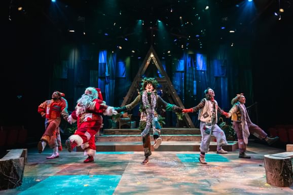 Five performers are hold hands and kicking out in a merry dance. They all wear warm-toned patchwork costumes. Confetti falls from above. Behind them is a wooden a-frame structure and a blue cloth backdrop.