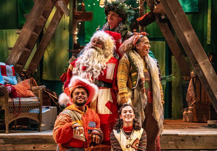Five performers are gathered in a staggered bunch over three steps. They all wear warm-toned patchwork costumes. They are all smiling, painting a very happy scene. Behind them is a wooden a-frame structure.