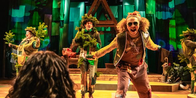 A young girl is sat with her back to the camera as a woman sings at her with a passionate expression. There are three other performers on the stage, who are all dressed as trees in green patchwork outfits complete with branches in their hands and on their heads. Behind them is a wooden a-frame structure and a green cloth backdrop.