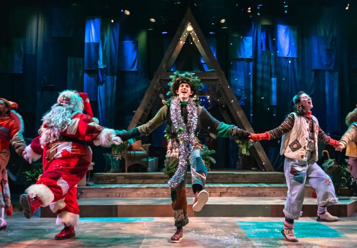 Five performers are hold hands and kicking out in a merry dance. They all wear warm-toned patchwork costumes. Confetti falls from above. Behind them is a wooden a-frame structure and a blue cloth backdrop.