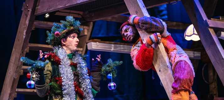 Two performers are talking, the one on the left is dressed as a tinsel covered tree and the other as a cat. Both of their costumes have an earthy, patchwork quality. The performer dressed as a cat is wrapped around a wooden framed structure. The cat looks questioning while the tree looks clueless.