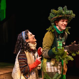 A young girl stands next to another performer, who is dressed as a tree. She wears a stripey jumper and a white gilet, embellished with patches. The other performer wears green patchwork overalls, a pine wreath on their head and hold branches in their hands. They share a sweet moment, both smiling.