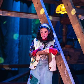 A young girl leans against a wooden framed structure with a sketchbook in her hand. She wears denim dungarees, a stripey jumper and a white gilet, embellished with patches. She smiles into the distance as she draws in the book.