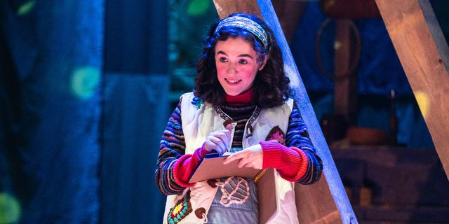 A young girl leans against a wooden framed structure with a sketchbook in her hand. She wears denim dungarees, a stripey jumper and a white gilet, embellished with patches. She smiles into the distance as she draws in the book.