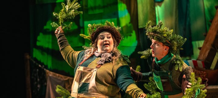 Two performers are dressed as Christmas trees in green patchwork overalls. They wear pine wreaths on their heads and hold branches in their hands. Behind them is a patchwork green cloth backdrop. Their expressions are apprehensive.