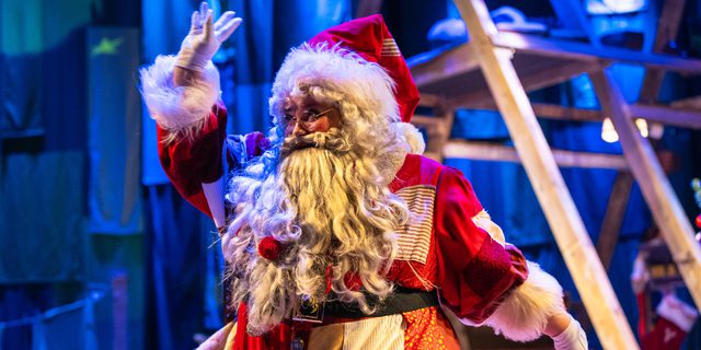 A performer dressed as Father Christmas in a red and white patchwork costume is waving. They wear a big white beard and white gloves. In the background there’s a wooden framed structure in a wash of blue light.