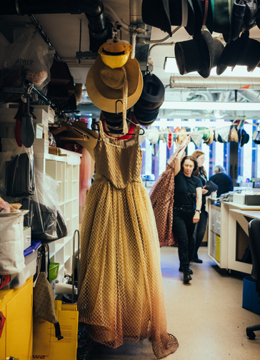 Image taken from the perspective of someone standing in the doorway of our wardrobe workshop, hats of all varieties hang from the ceiling and a dress is waiting to be steamed.