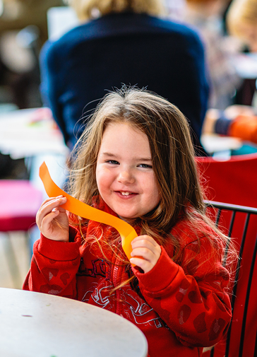 A young girl, holds a cut piece of orange paper in her hands whilst smiling at the camera.