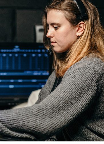 A young woman with light brown hair sits working on a technical programming desk, surrounded by screens