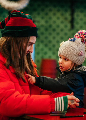 An elf (wearing a red puffer jacket and green and red pointy hat) chats with a little girl.