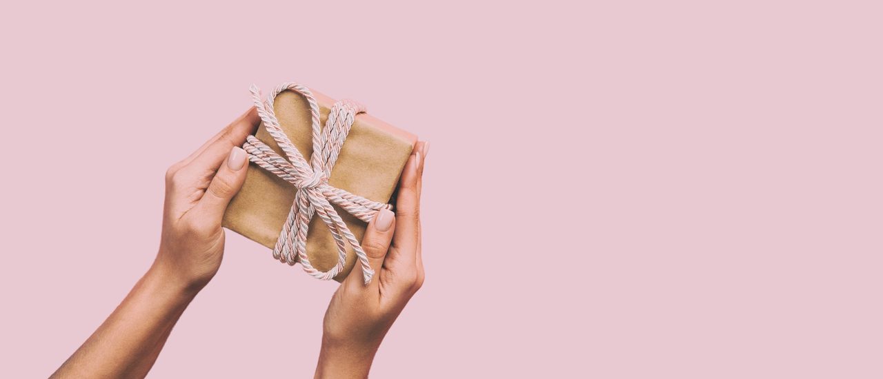 Two hands holding a brown paper package, tied up with string on a pastel pink background.
