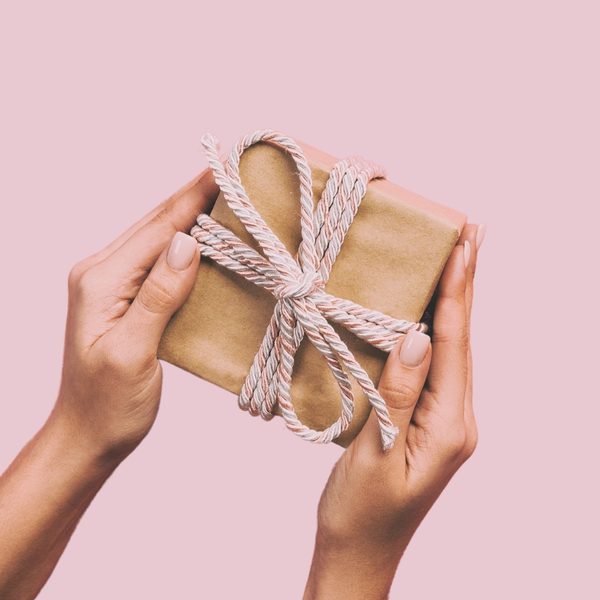Two hands holding a brown paper package, tied up with string on a pastel pink background.