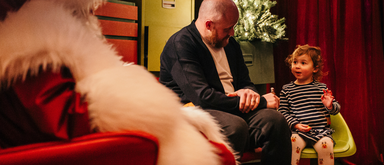 Father Christmas in a red suit with fluffy white trim sits in his grotto with a small child and father.