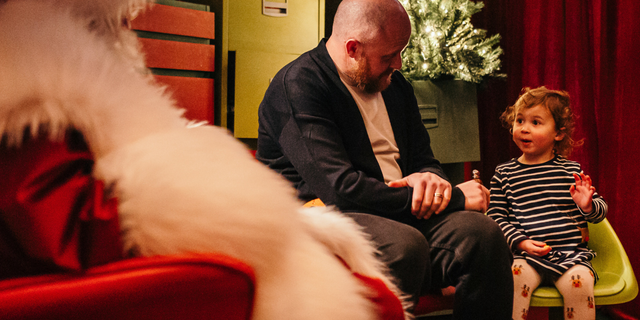 Father Christmas in a red suit with fluffy white trim sits in his grotto with a small child and father.