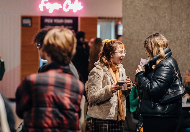 Two women stand in the Festival Theatre foyer, smiling at eachother. They are holding drinks and phones. In front of them are two other people with their faces obscured. In the background is a glowing pink sign which says 'Foyer Cafe'